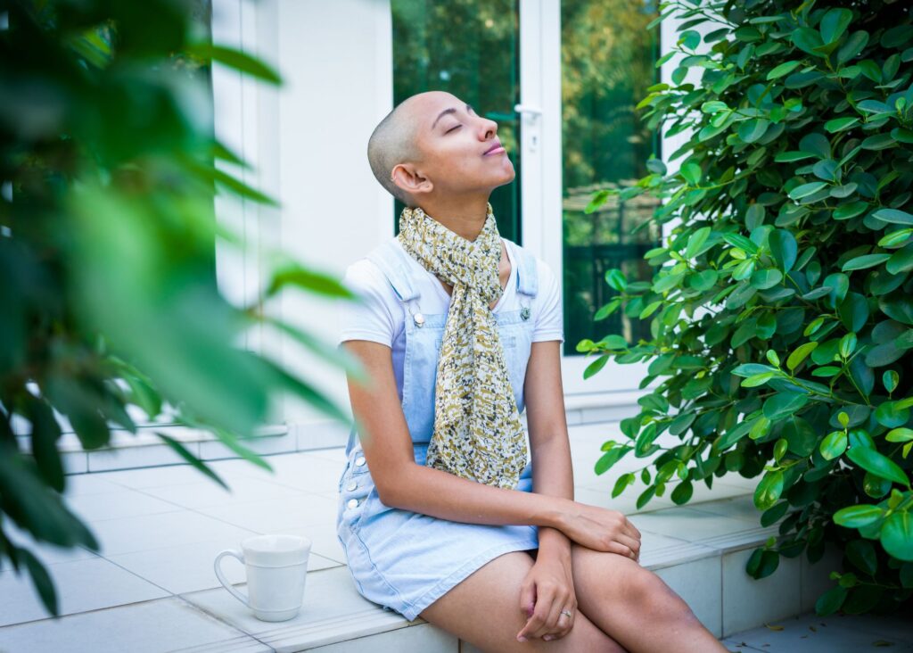 A an individual wearing a blue Dres and multi-colored scarf sitting outside on a step with their head tilted back and their eyes closed, as if to take in the day. They are surrounded by plants with a white coffee cup next to them.