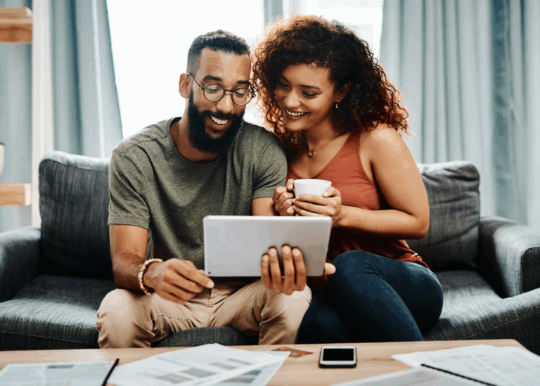 A woman and man sit on the couch and look at the scree of the tablet he is holding in his hands.