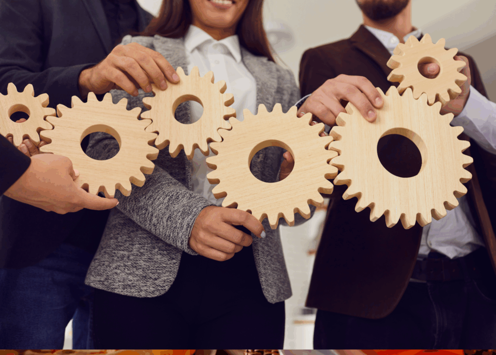 Several colleagues stand together as they each hold a wooden gear. Each gear is connected to another colleague's gear. Their faces are not visible. Just a closeup of all the wooden gears interconnected.